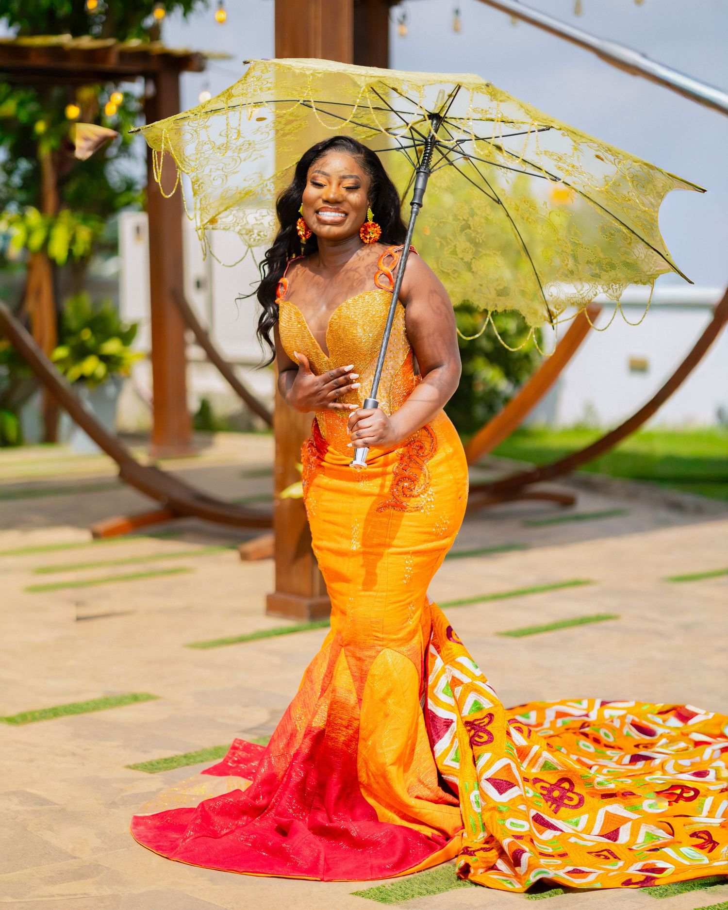 A vibrant orange and red dress flows gracefully as someone poses with a yellow umbrella in an outdoor garden setting.