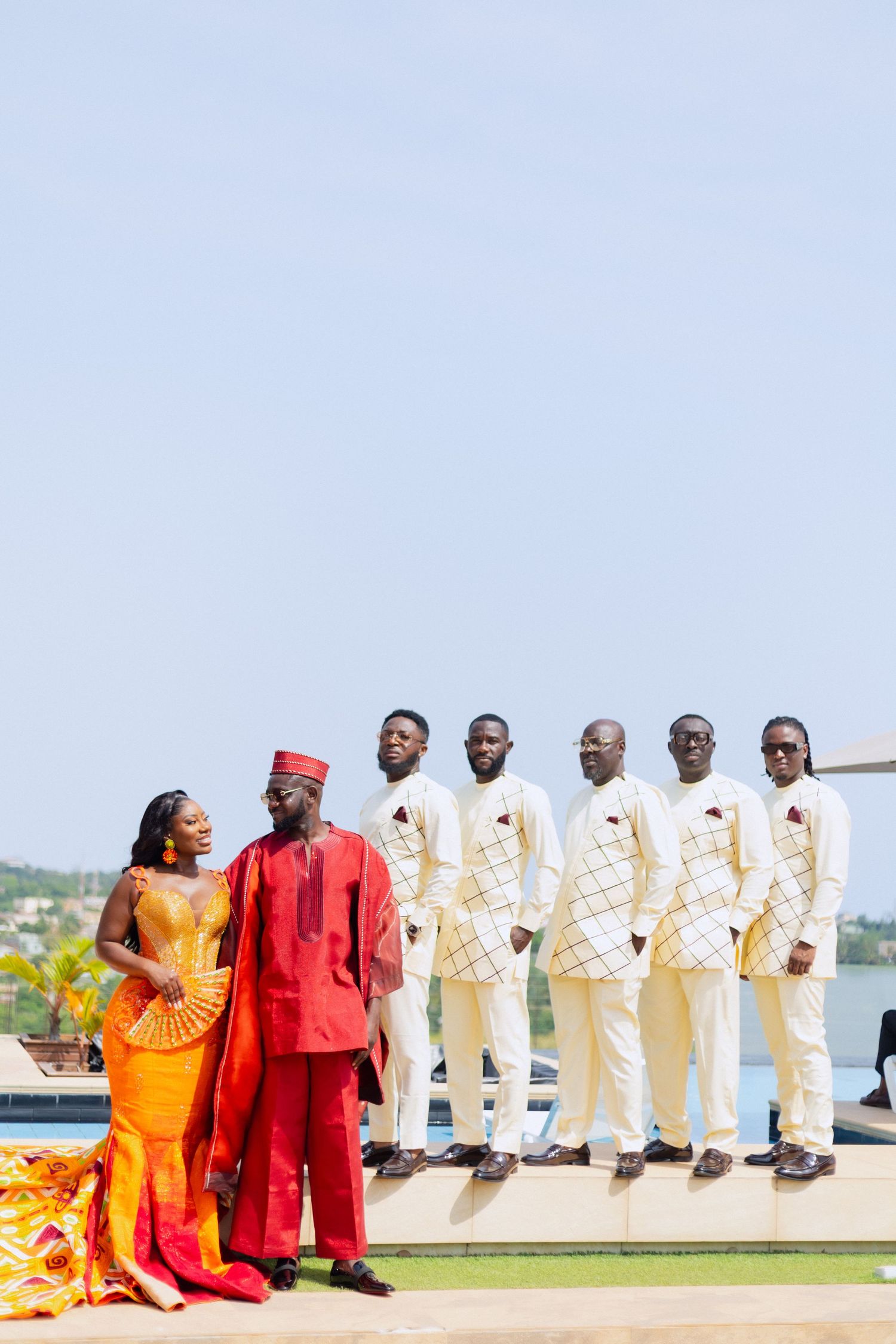 A wedding party poses on a waterfront with groomsmen in white suits and a couple in vibrant red and orange attire.