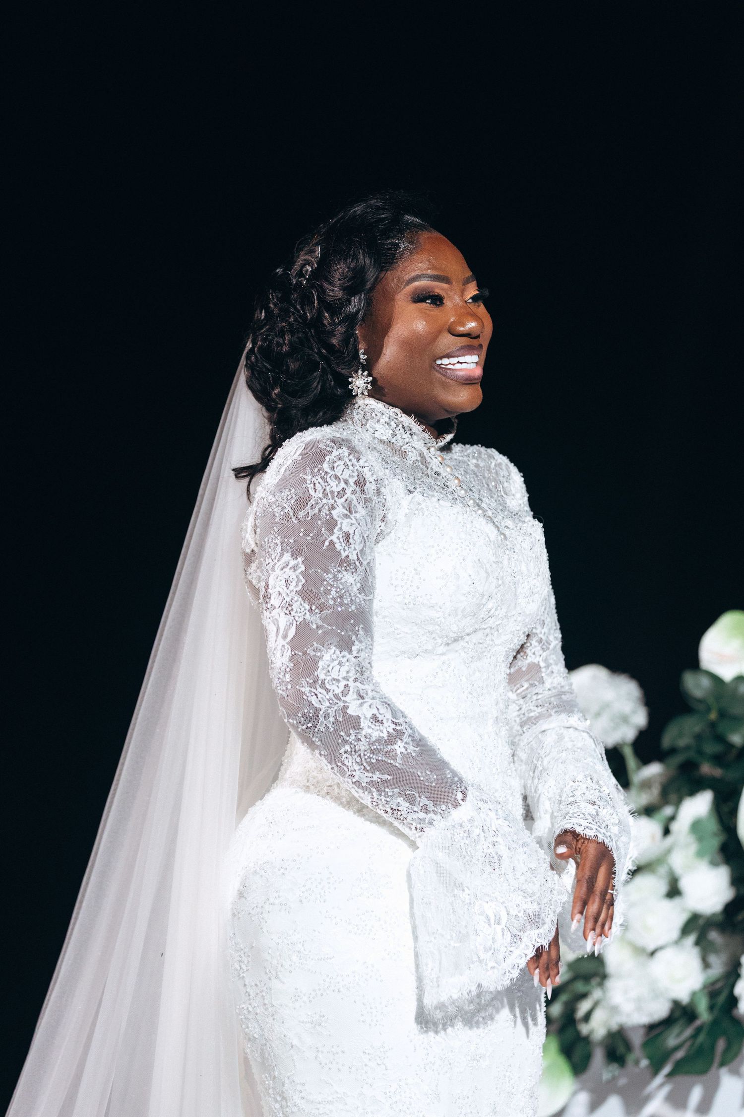 A bride in an elegant long-sleeved white lace wedding gown and veil poses against a dark background with white flowers.