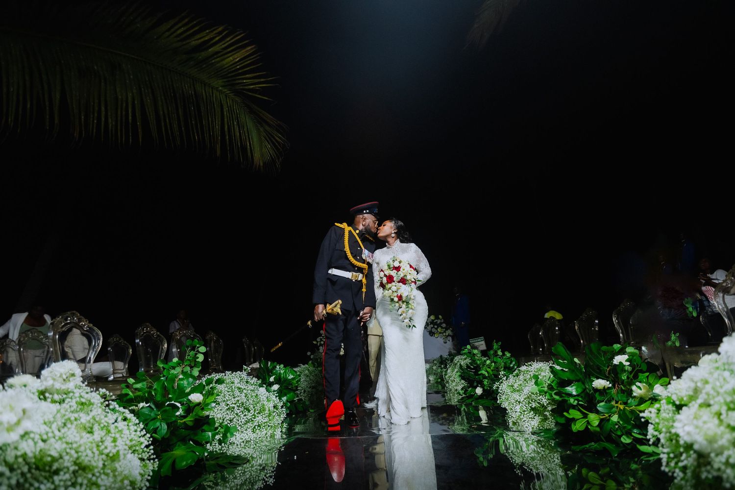 A couple dances together at an outdoor wedding reception surrounded by lush greenery and white flowers in the evening.