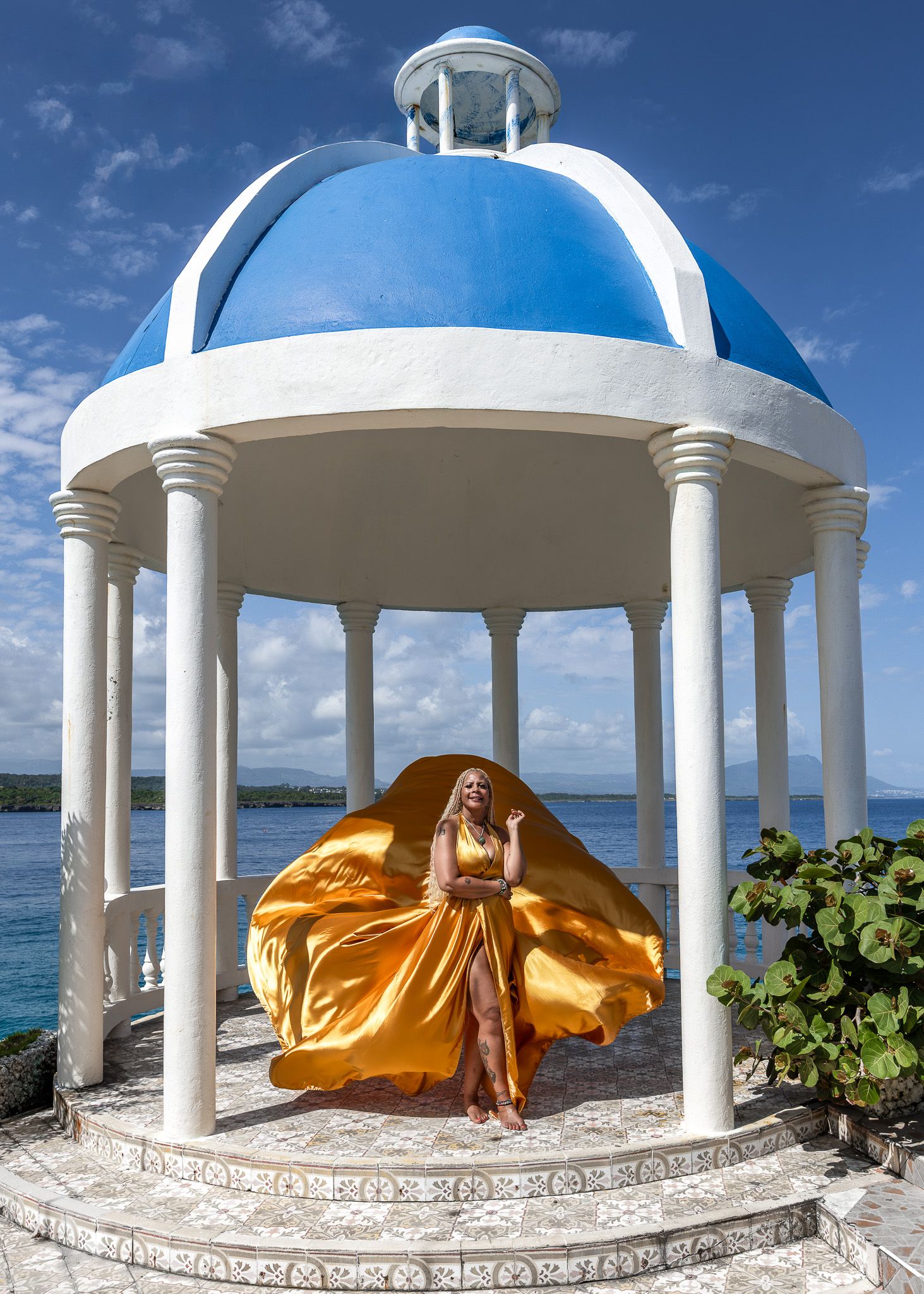 Dramatic fashion photo of flowing gold dress in white domed gazebo by ocean.
