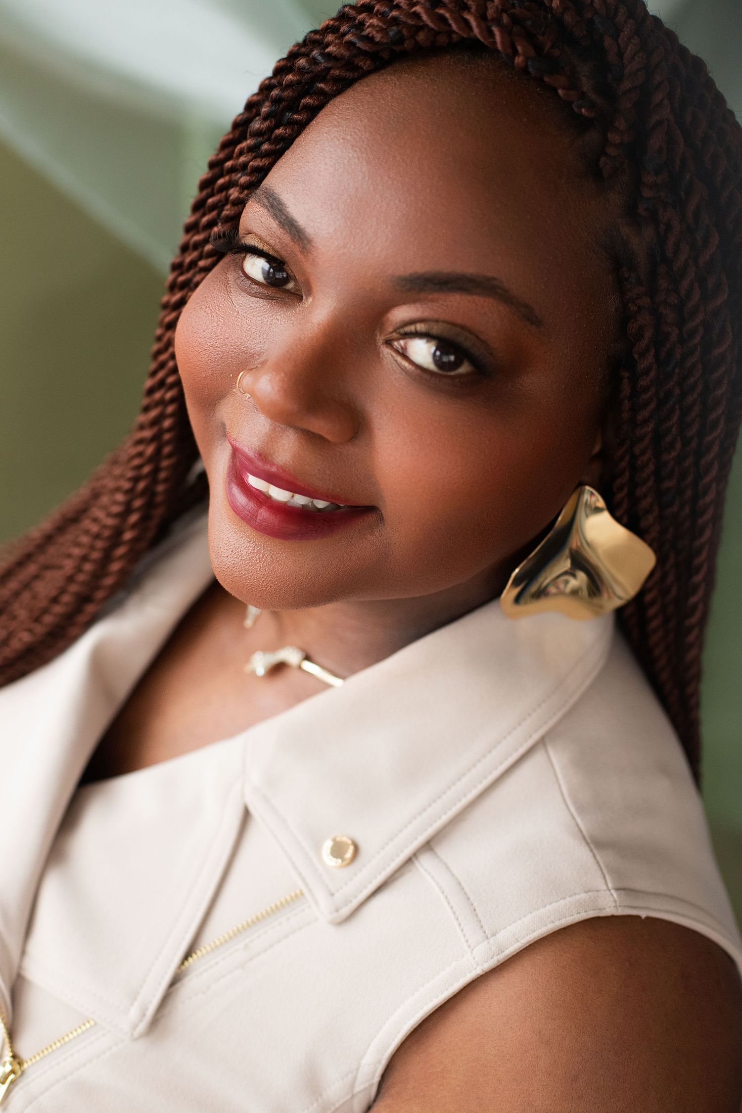 Professional portrait showing warm smile and reddish-brown braids, wearing an elegant cream-colored sleeveless top.
