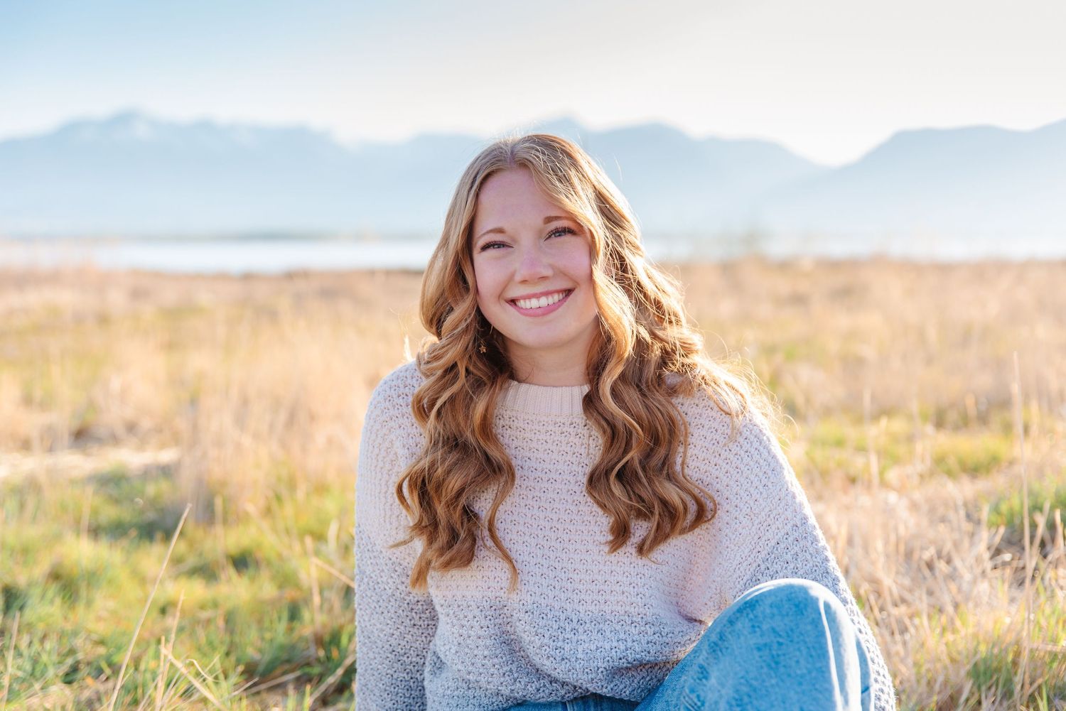 Woman in cozy sweater sits outdoors in golden field with mountains in distance.