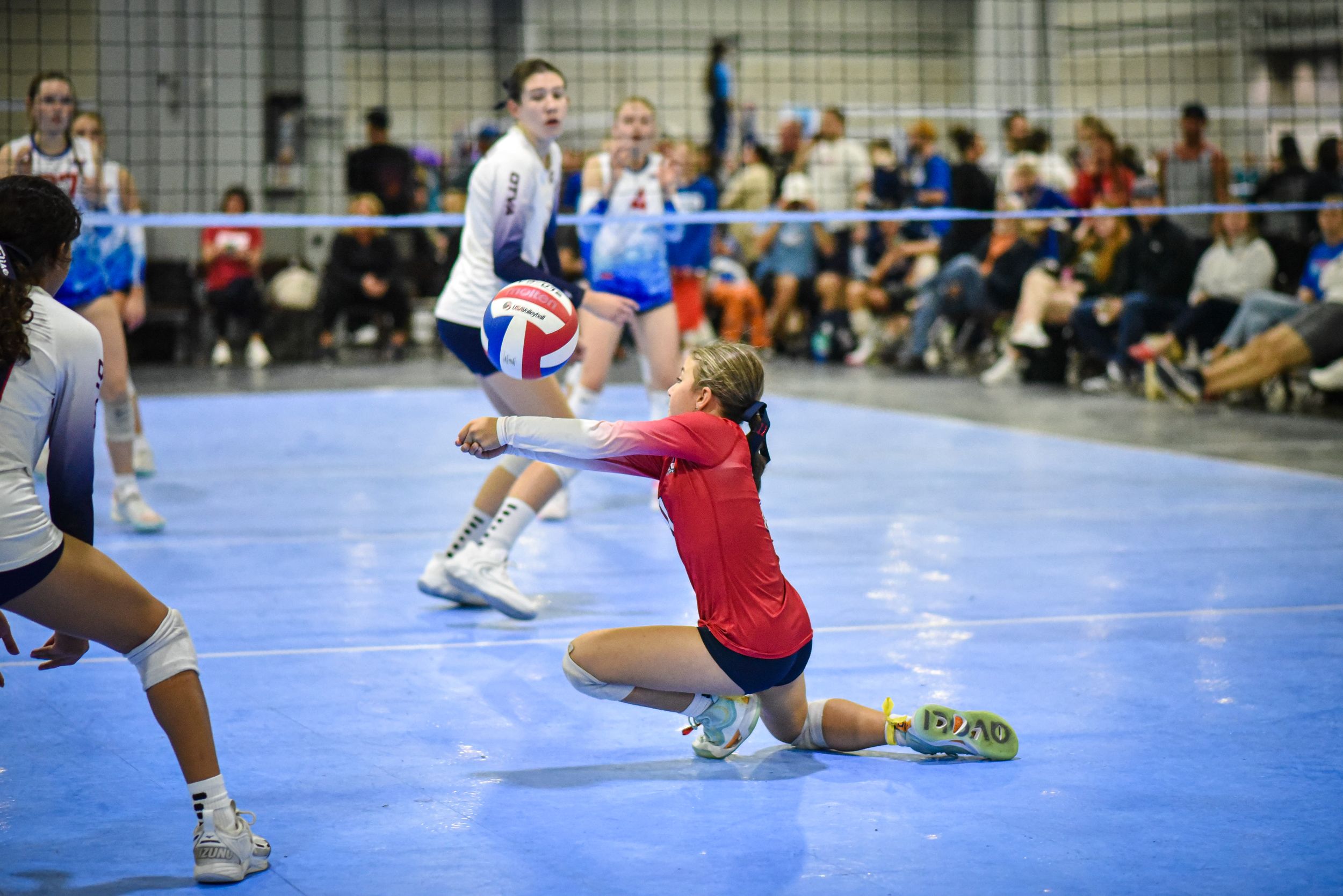 Female volleyball player in red jersey diving to dig the ball during an indoor competitive match.