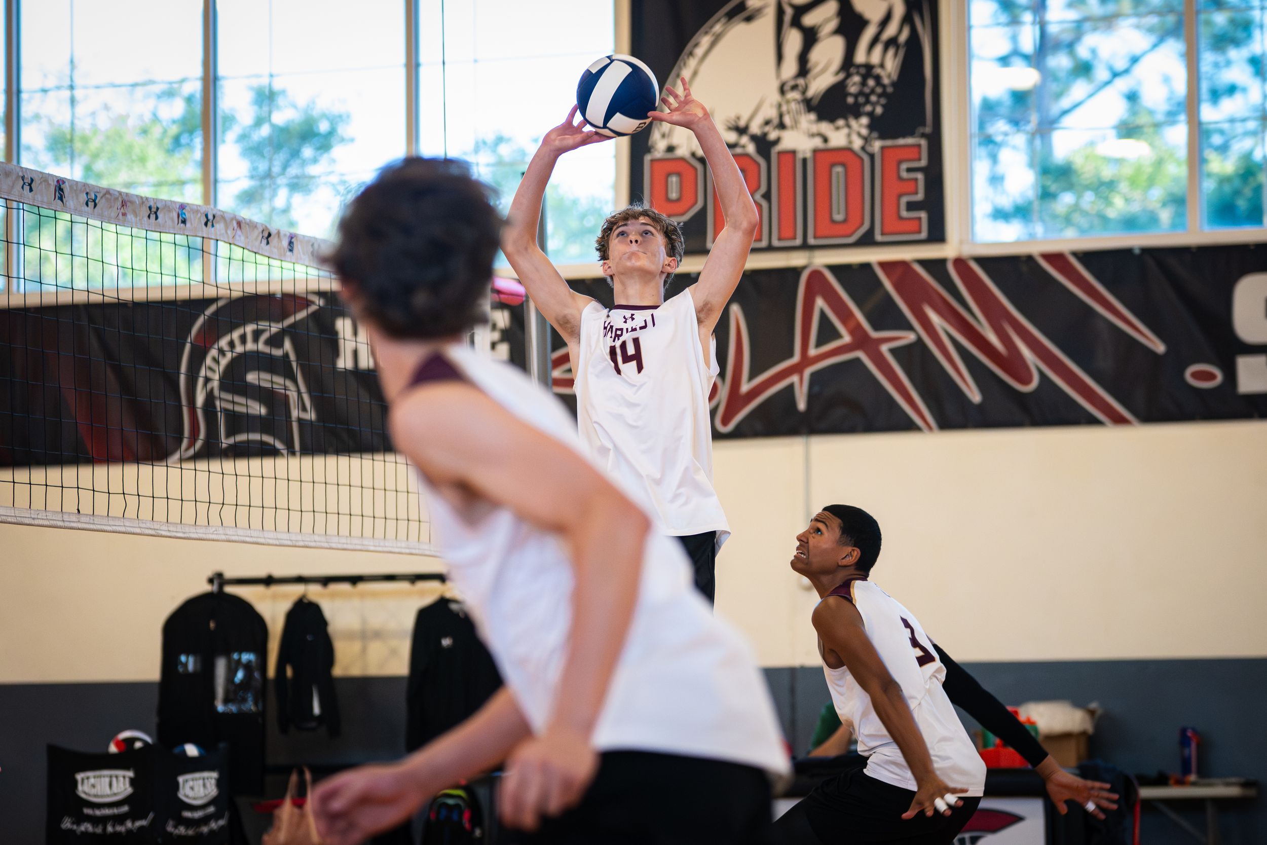 Male volleyball player #14 sets the ball during an indoor high school volleyball game in a gym.