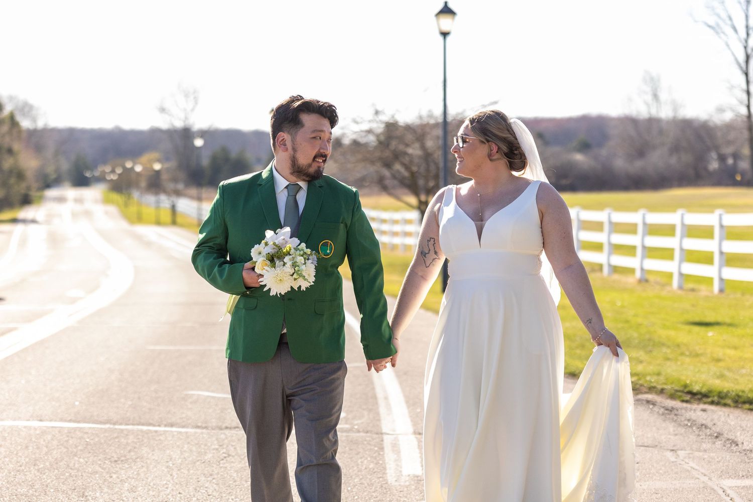 A bride and groom walk hand in hand along a sunny path beside white fenced fields.
