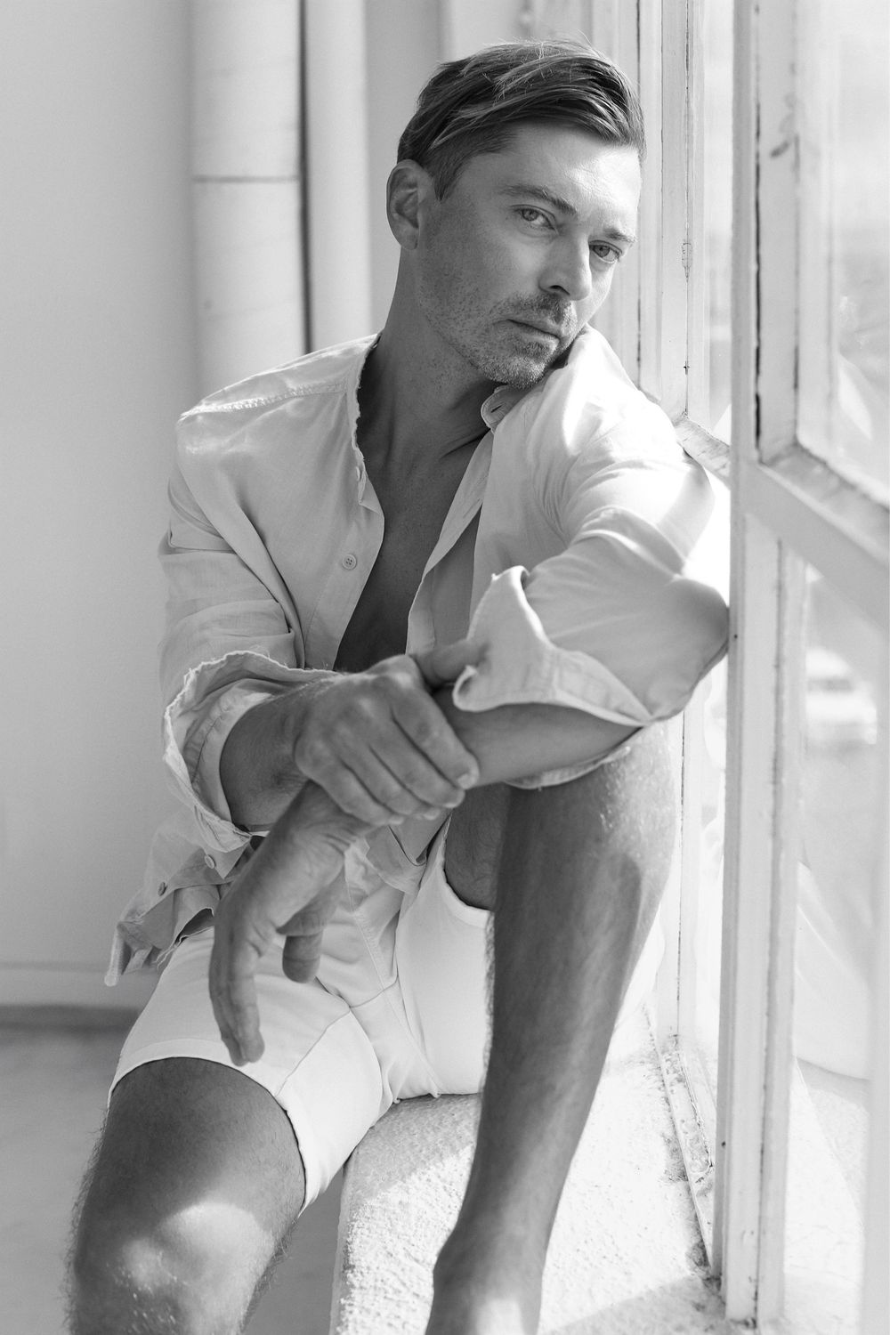 Black and white portrait of man in white shirt sitting by window.