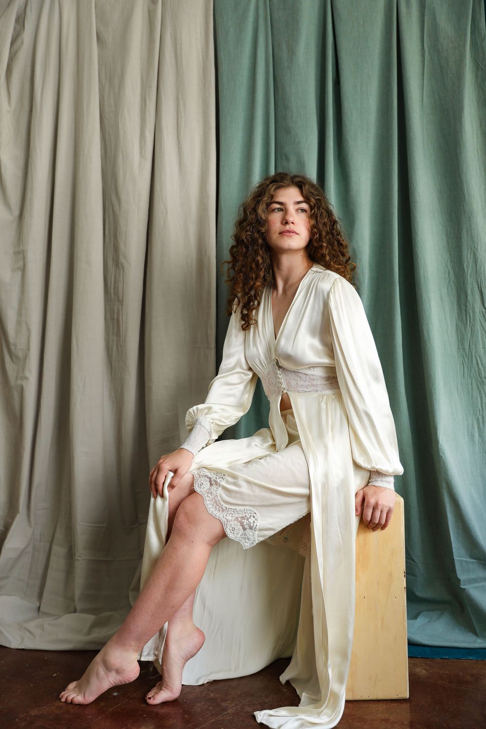 Woman in ivory satin robe with lace trim sits barefoot on white cube against sage green draped fabric backdrop.