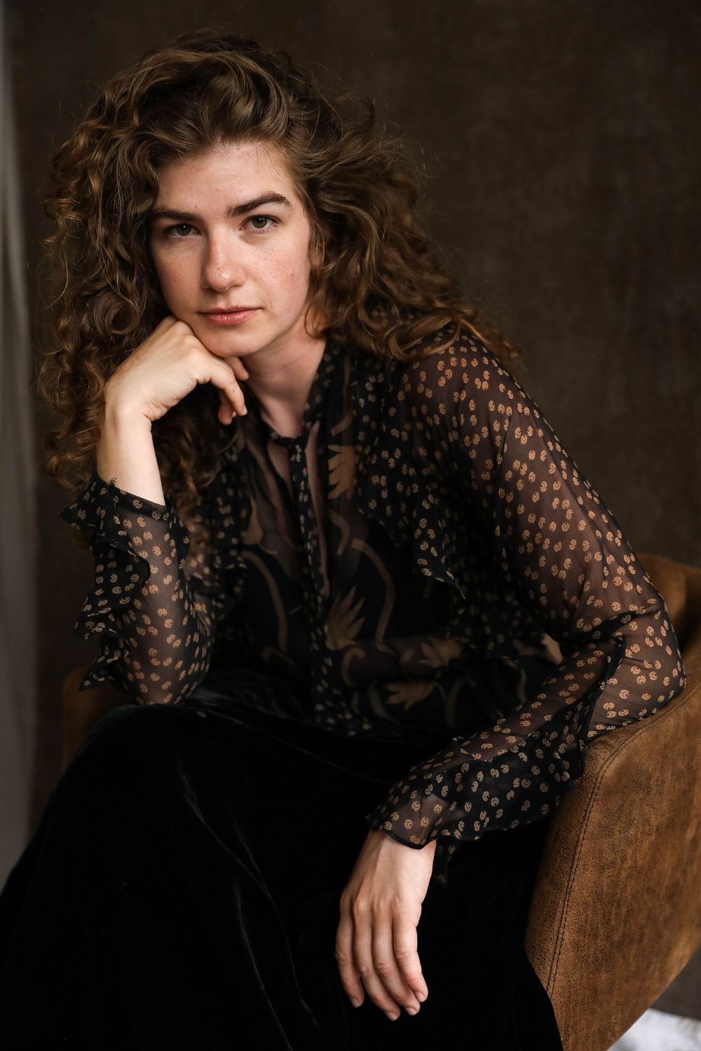 Young woman with curly brown hair in sheer black polka dot blouse sits thoughtfully in leather chair.