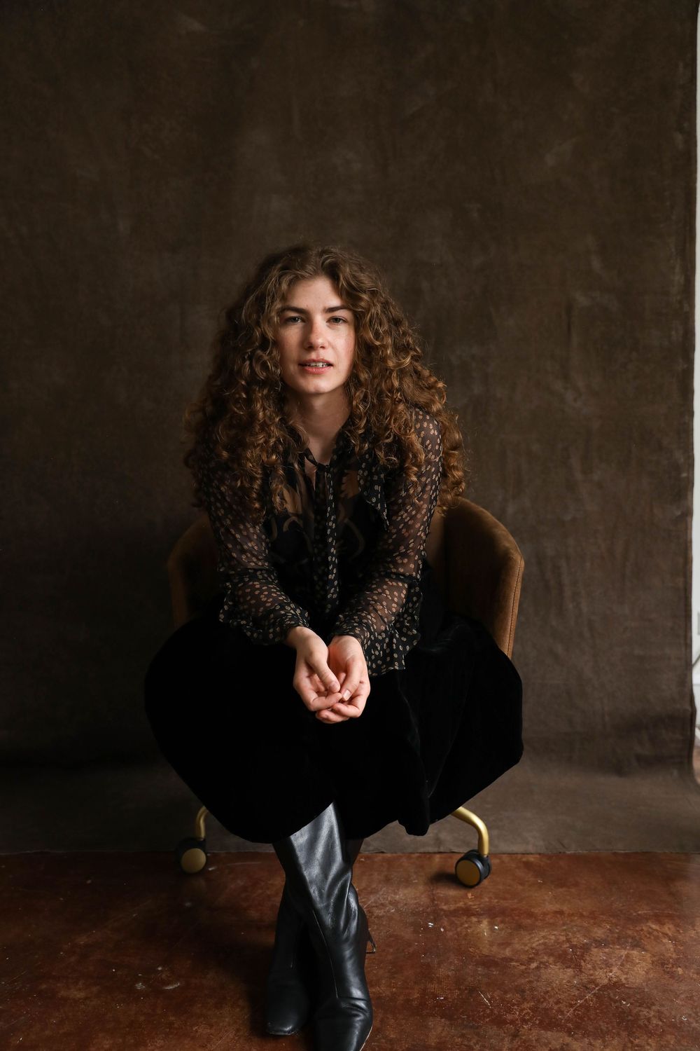 Young woman with curly hair sitting in a velvet chair, wearing black outfit and boots against a gray wall.
