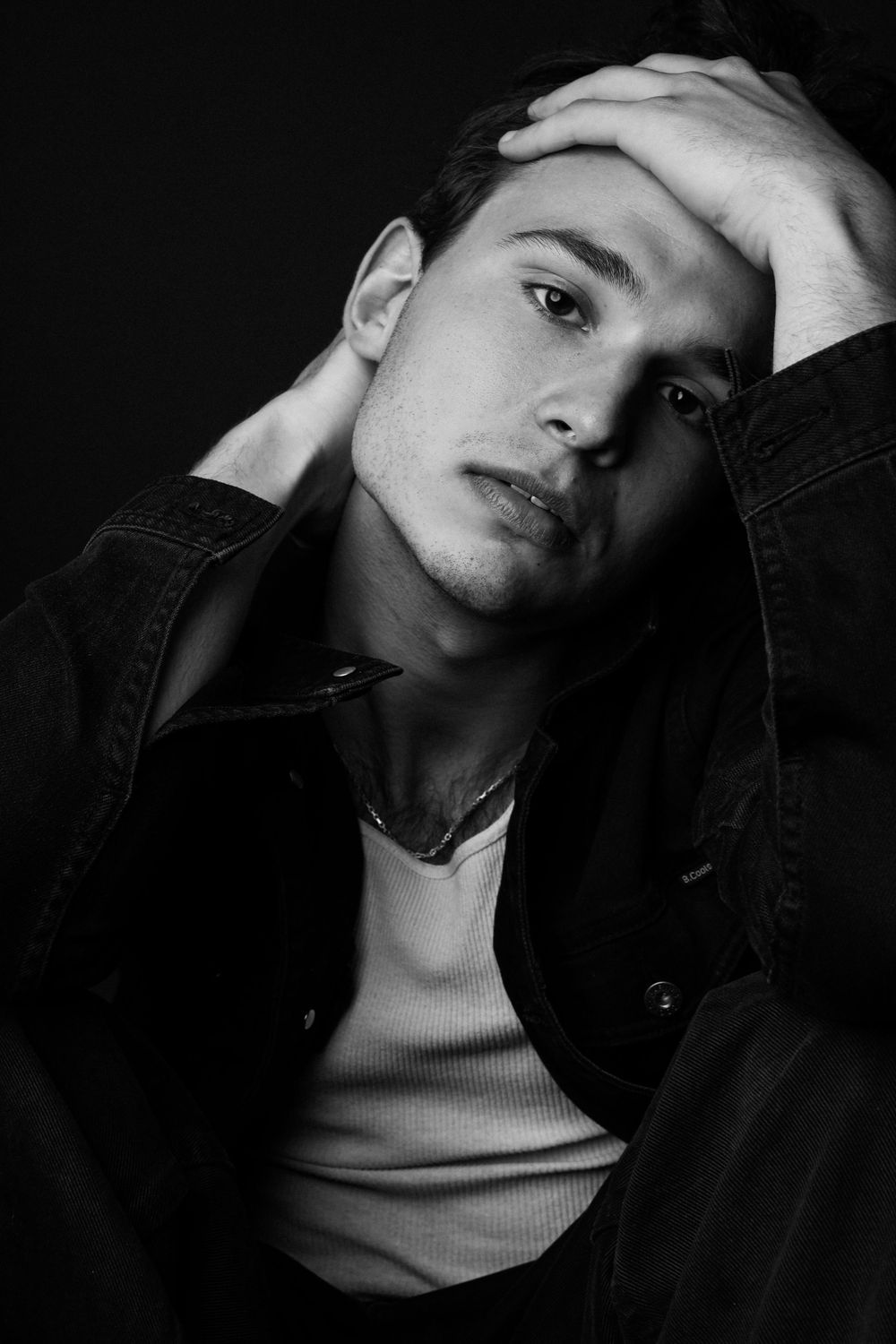 Black and white portrait of a young man in a denim jacket, hands on head, gazing pensively against dark background.