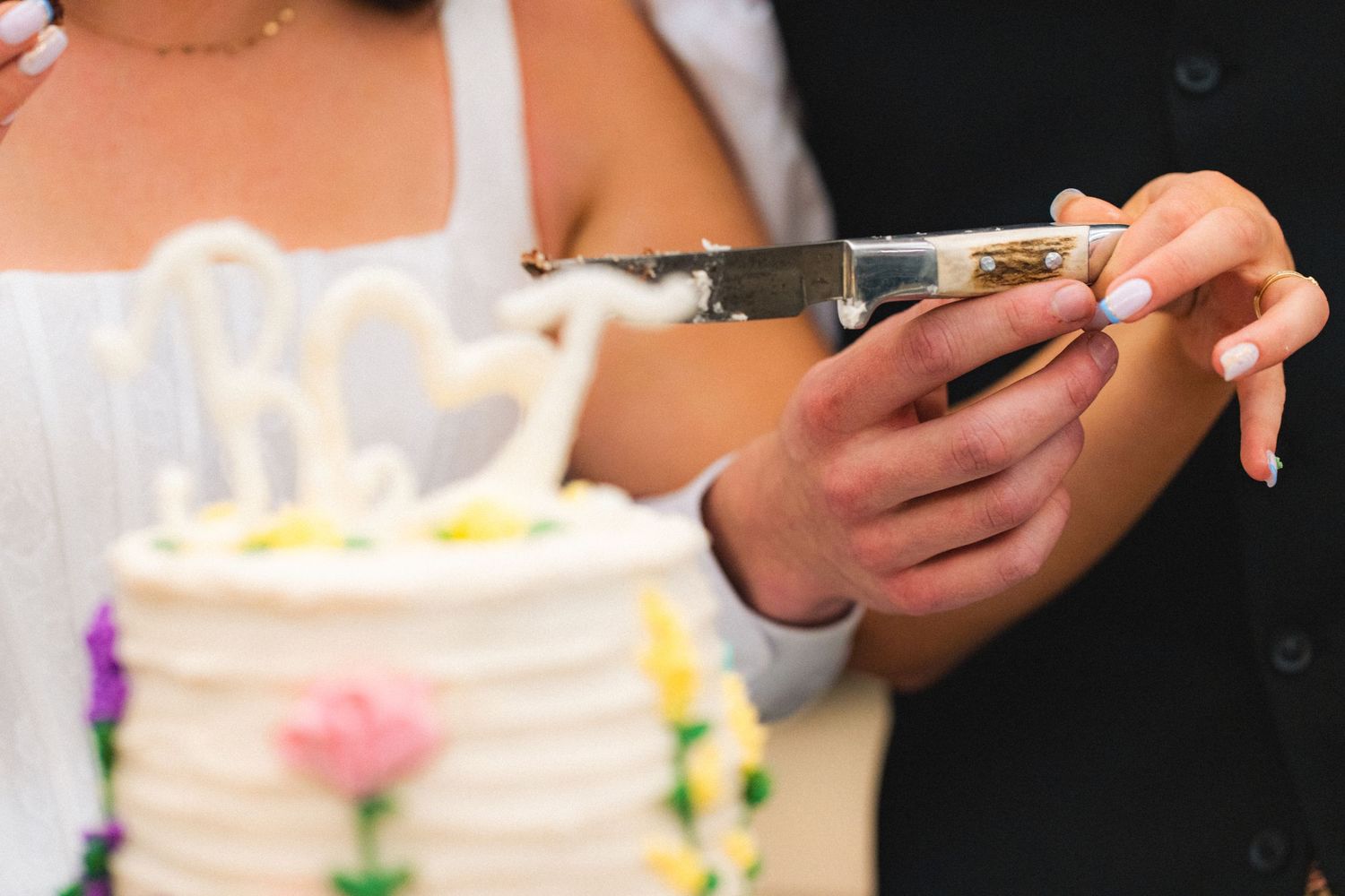 Couple holding knife together to cut elegant white wedding cake with colorful flowers.