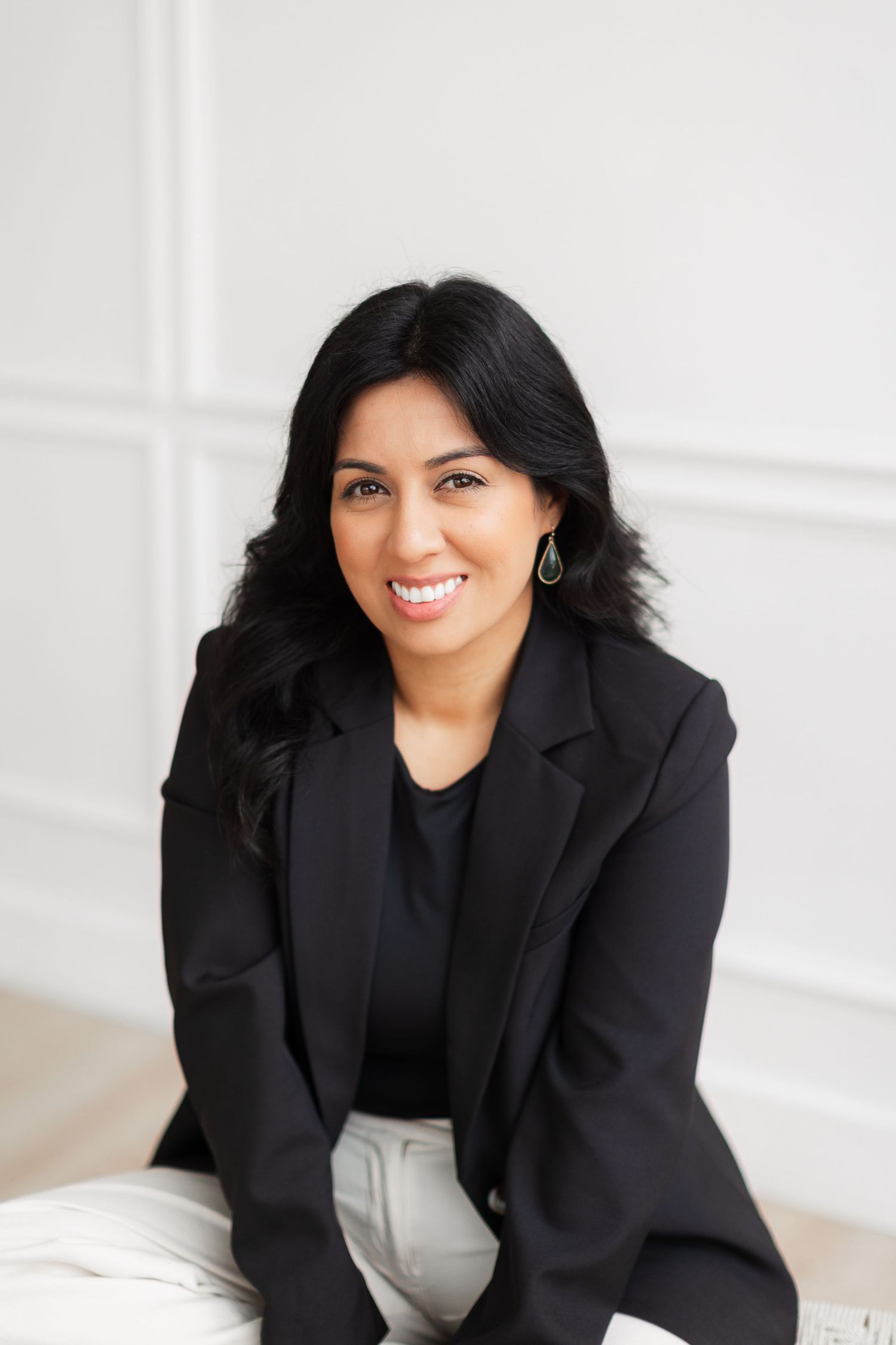 Professional woman wearing black outfit sits in tan leather chair against white wall.