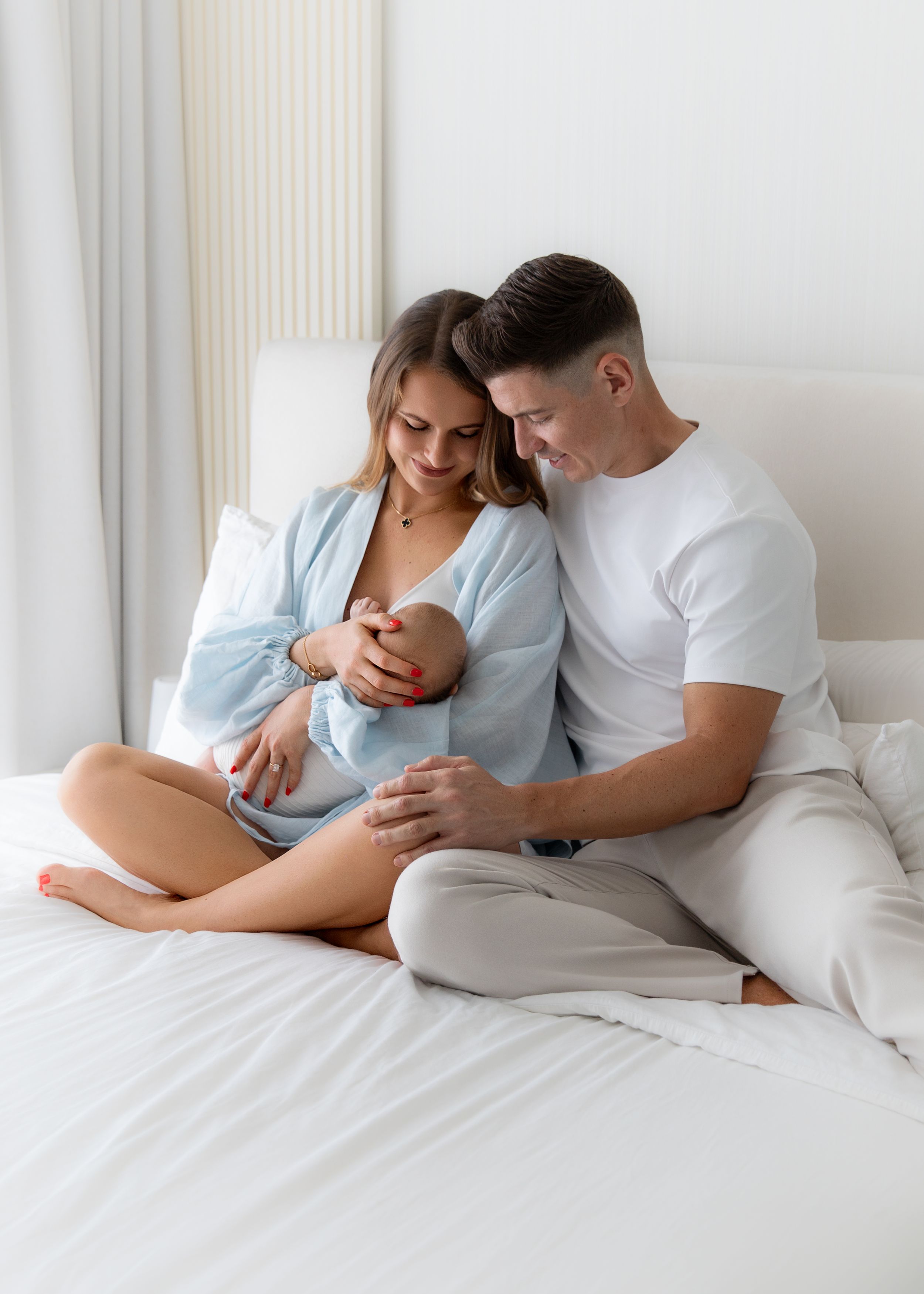 Mum, dad and newborn baby sitting on the bed with a white background. baby in mums arms. mum and dad looking at baby