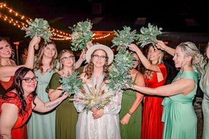 Wedding party in colorful dresses hold green floral bouquets under string lights during evening celebration.