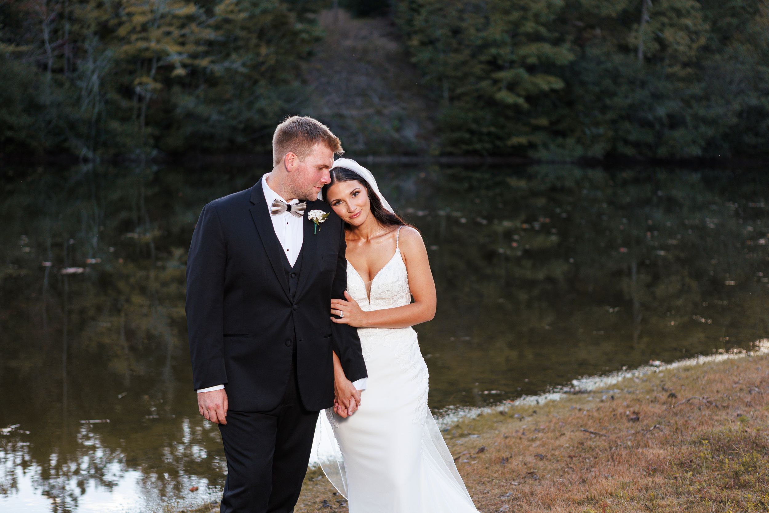 Couple embracing by a serene lake with trees reflecting on the water's surface.