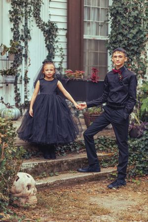 Playful siblings in matching black outfits pose together in front of an ivy-covered house exterior.
