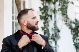 A person in a black jacket poses thoughtfully against a white wall with green foliage in the background.