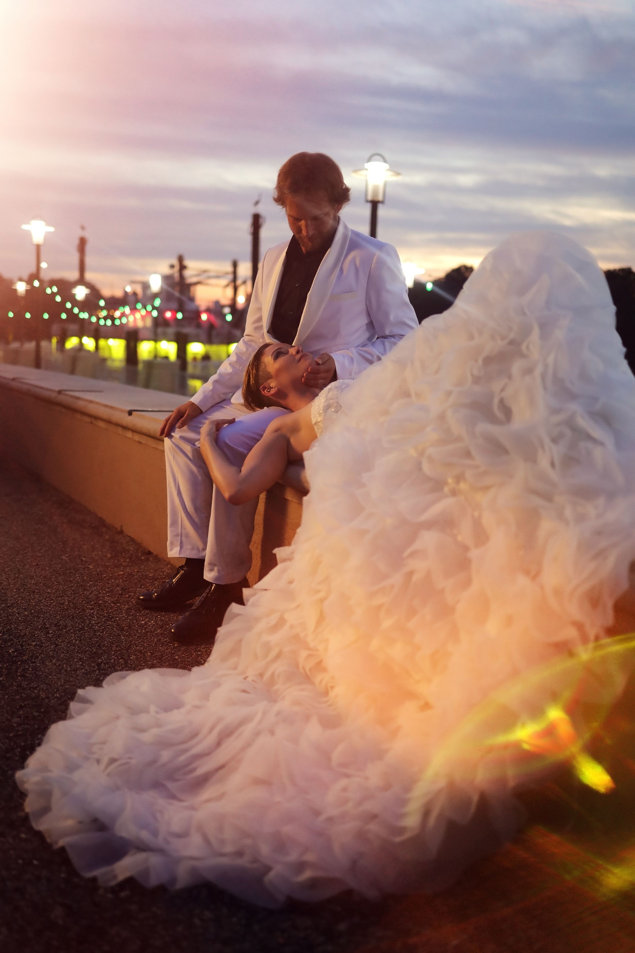 A couple sits together at sunset on a boardwalk with string lights and a flowing white wedding dress.