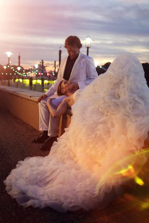 A couple sits together at sunset on a boardwalk with string lights and a flowing white wedding dress.