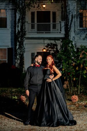 A romantic couple in formal black attire poses for Halloween-themed photos with pumpkins and outdoor lighting at dusk.