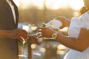 A wedding couple holds champagne glasses together in warm golden sunlight during an outdoor reception celebration.