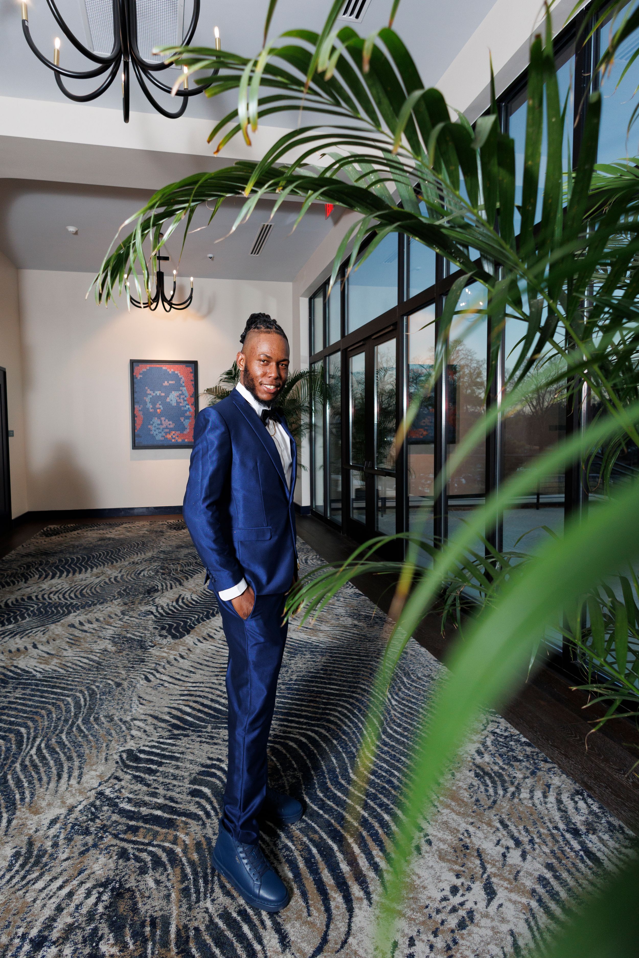 Professional in navy blue suit poses in elegant lobby with tropical plants and wavy patterned carpet.