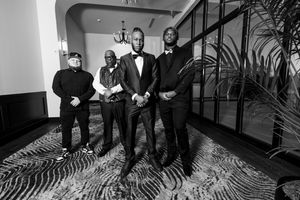 A black and white photograph shows a group of five people in formal attire standing on a patterned carpet.