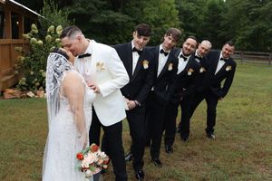 Wedding party in coordinated black suits stands in playful lineup formation outdoors while couple shares romantic moment.