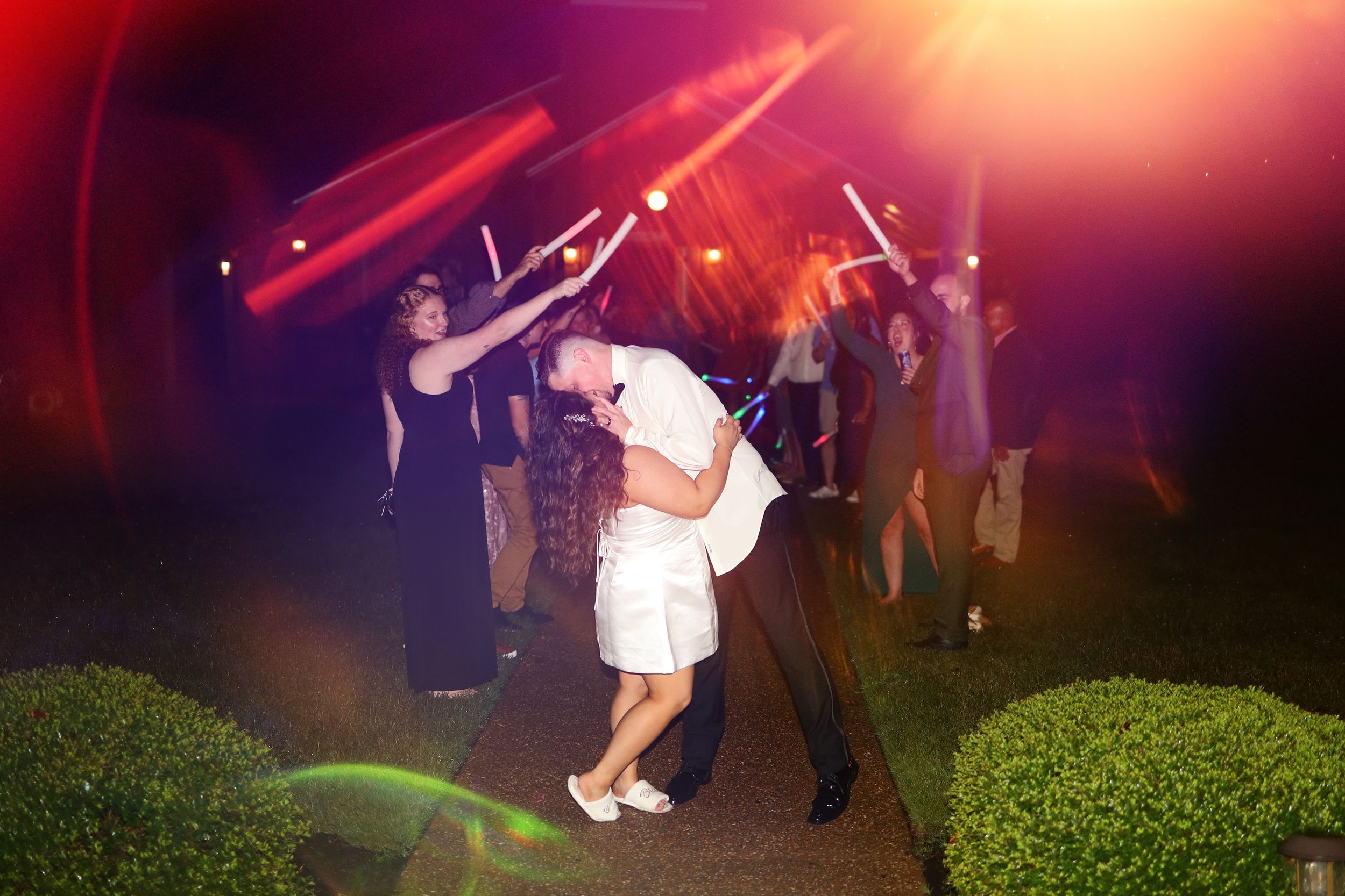 Wedding guests dance and celebrate on a dark outdoor patio illuminated by dramatic red lighting and lens flares.