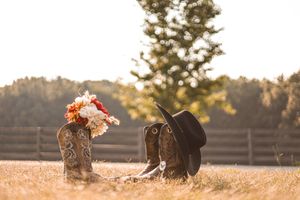 Two ornate cowboy boots with a black hat sit in golden sunlit grass with a wooden fence in the background.