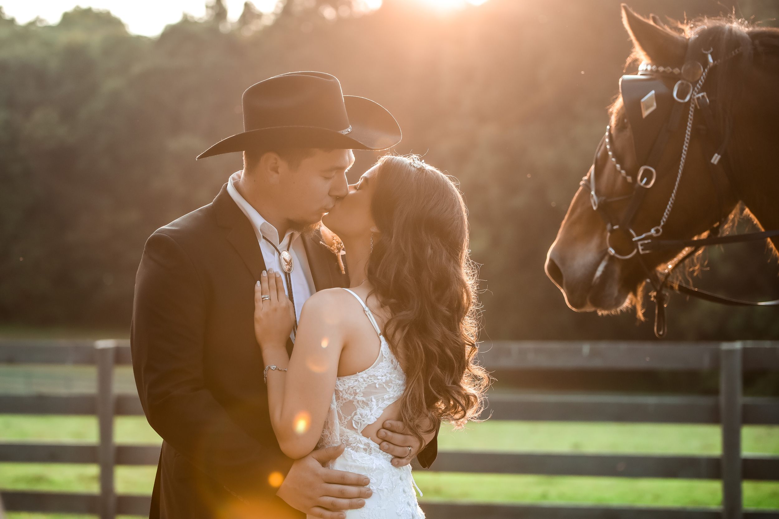 Romantic wedding couple embraces at sunset near horse stables with golden sunlight streaming through the trees.
