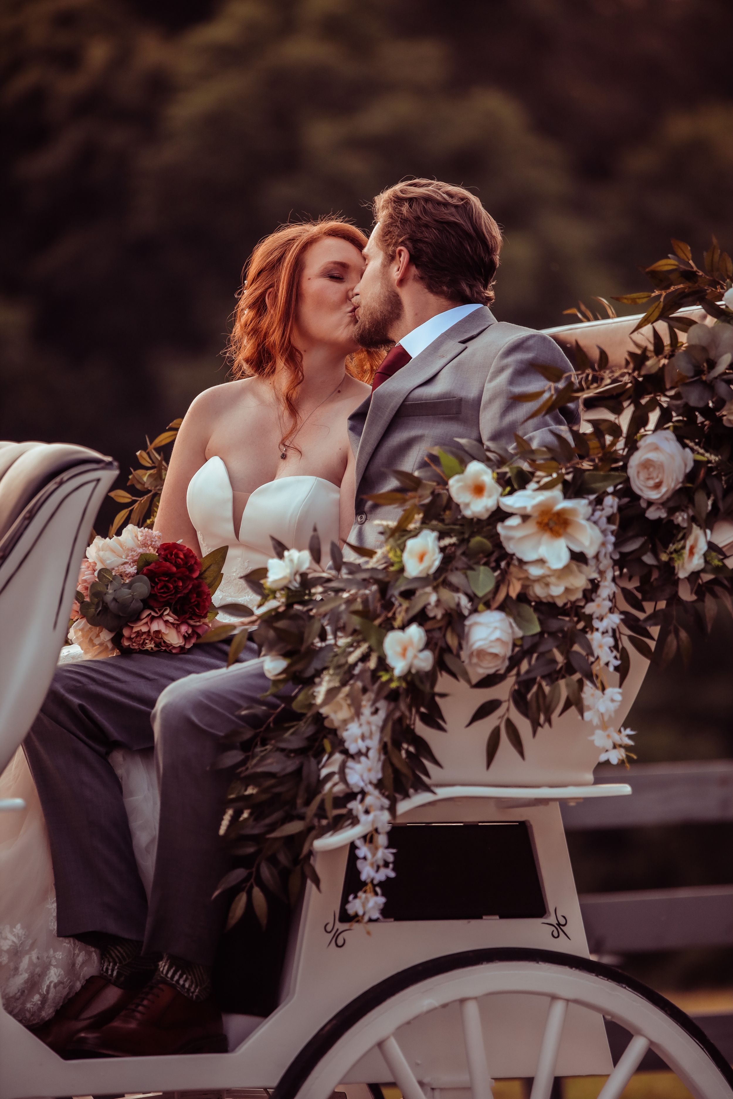 A romantic photo of a couple sharing a kiss while seated in a white horse-drawn carriage adorned with flowers.