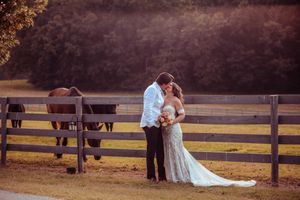 A romantic photo of a couple embracing near a wooden fence with horses grazing in the field during sunset.