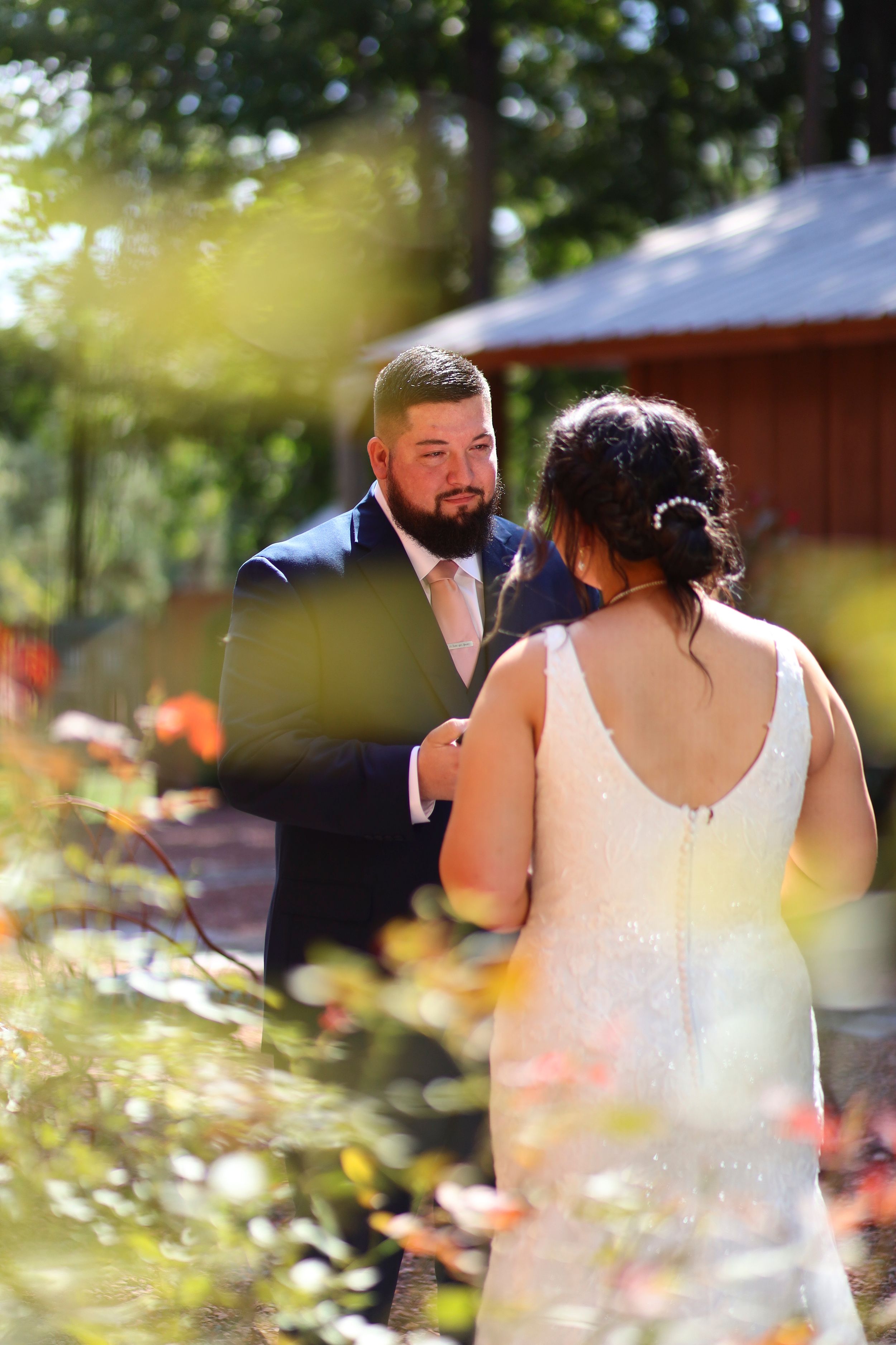 A couple shares an intimate moment during their outdoor wedding ceremony, with sunlit foliage and a rustic barn in the background.