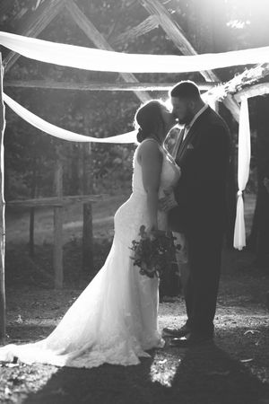 Black and white photo of a romantic wedding moment under draped fabric with sunlight streaming through trees in background.