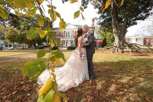 Two people in wedding attire embrace on a fall day outside a historic mansion with autumn leaves in the foreground.