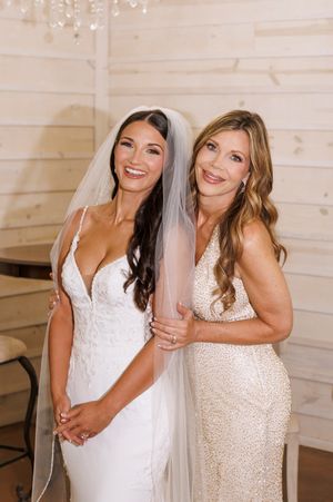 Two people in white dresses share joyful moments under crystal chandelier in rustic venue.