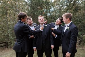 Groomsmen in black suits laughing together outdoors in a wooded setting.
