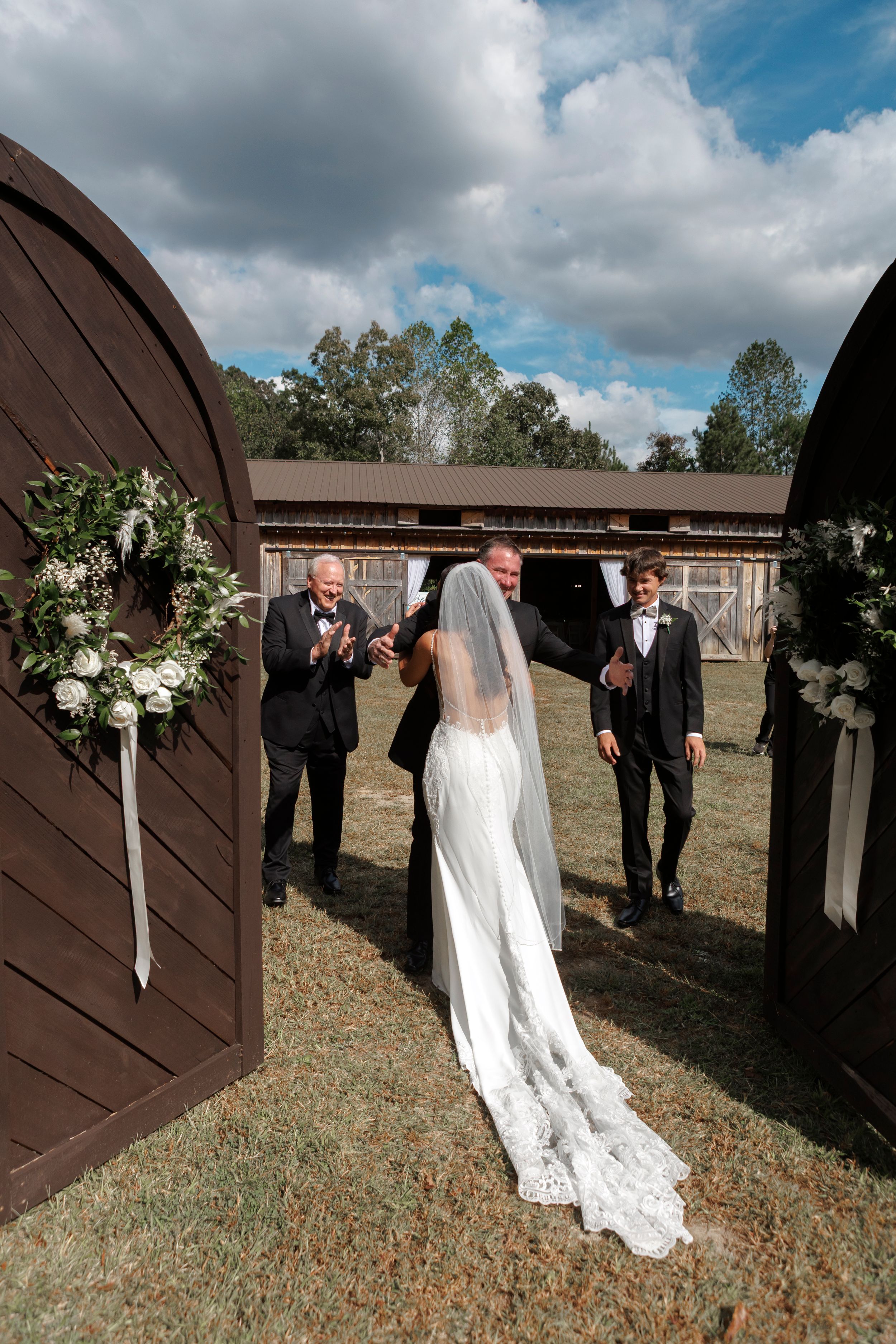 Wedding party poses in front of rustic brown barn doors on a grassy field under cloudy skies.