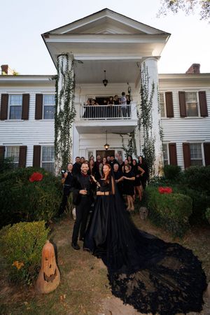 A group in black formal attire poses on the front porch of a white colonial-style house with Halloween decorations.