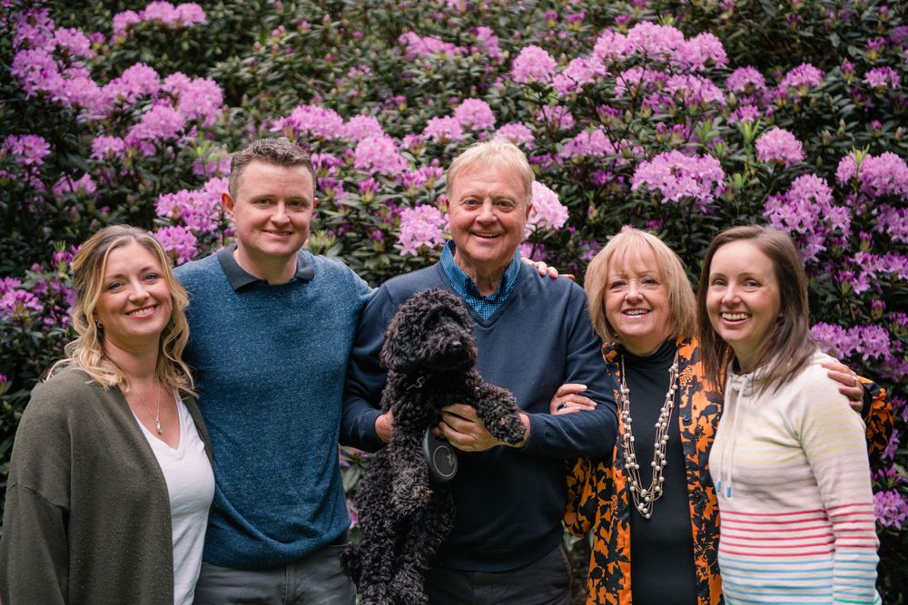 A group poses together outdoors in front of blooming pink rhododendron bushes with a black dog.