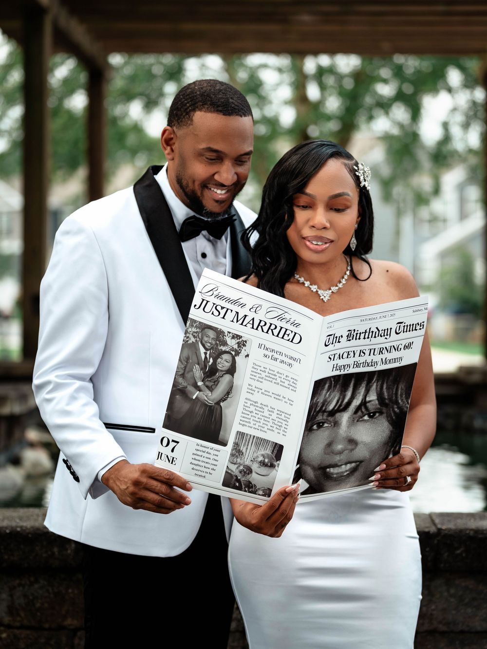 A couple in wedding attire share a joyful moment while reading a newspaper together outdoors.