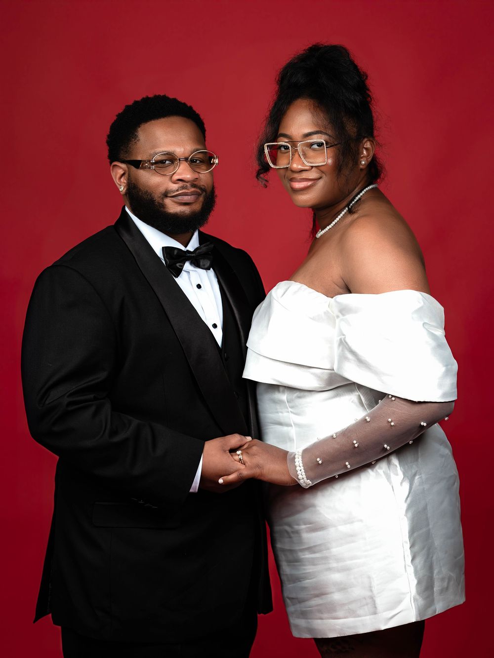 A couple in formal attire poses together against a red backdrop for an elegant portrait session.