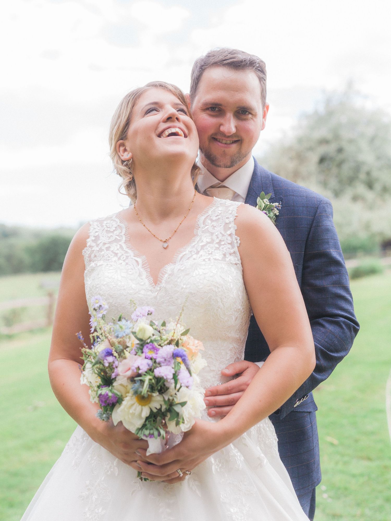 Joyful wedding couple sharing a candid moment outdoors with a purple and white floral bouquet.