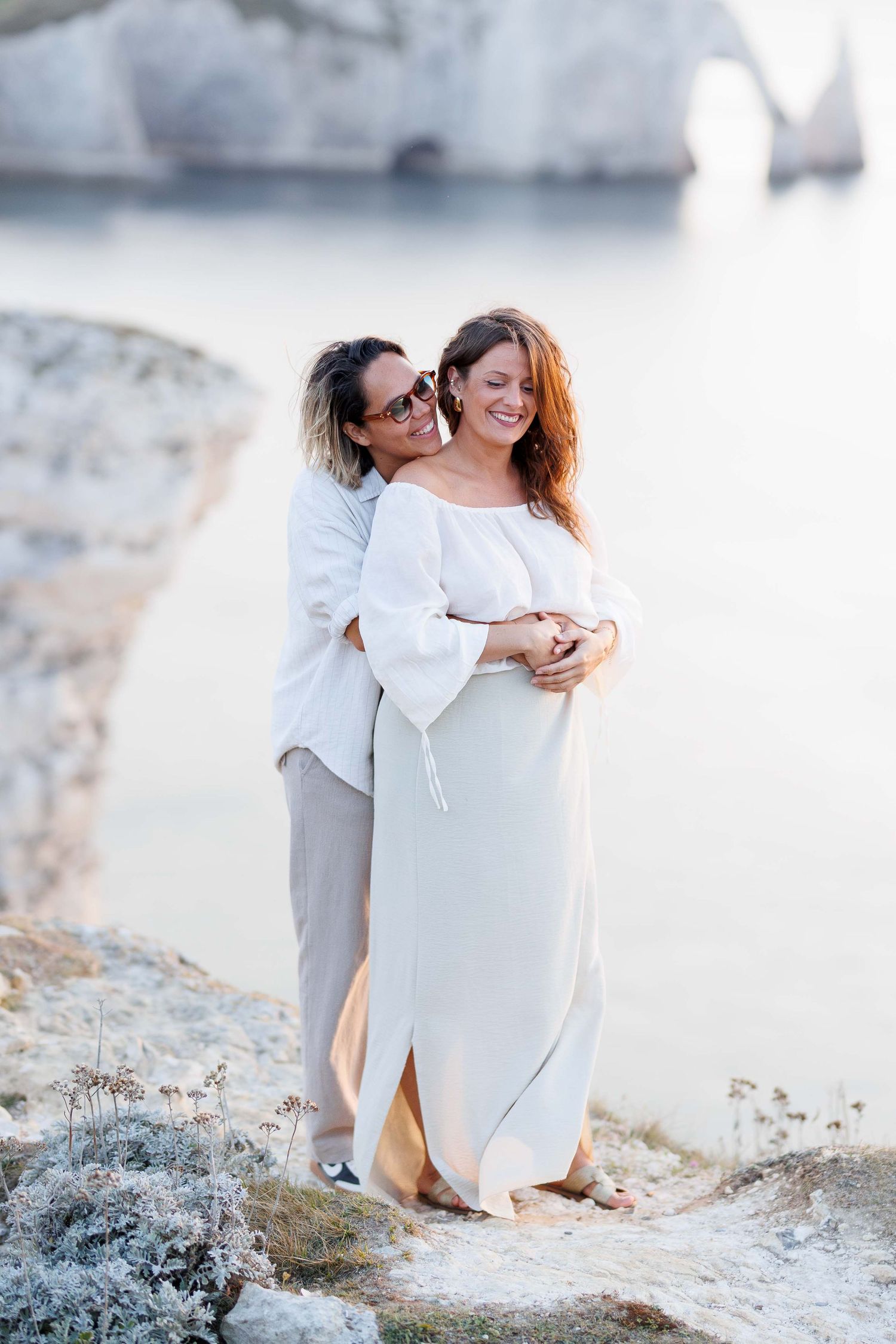 Two women in white outfits embrace on coastal cliffs with scenic sea arch and calm waters in background.