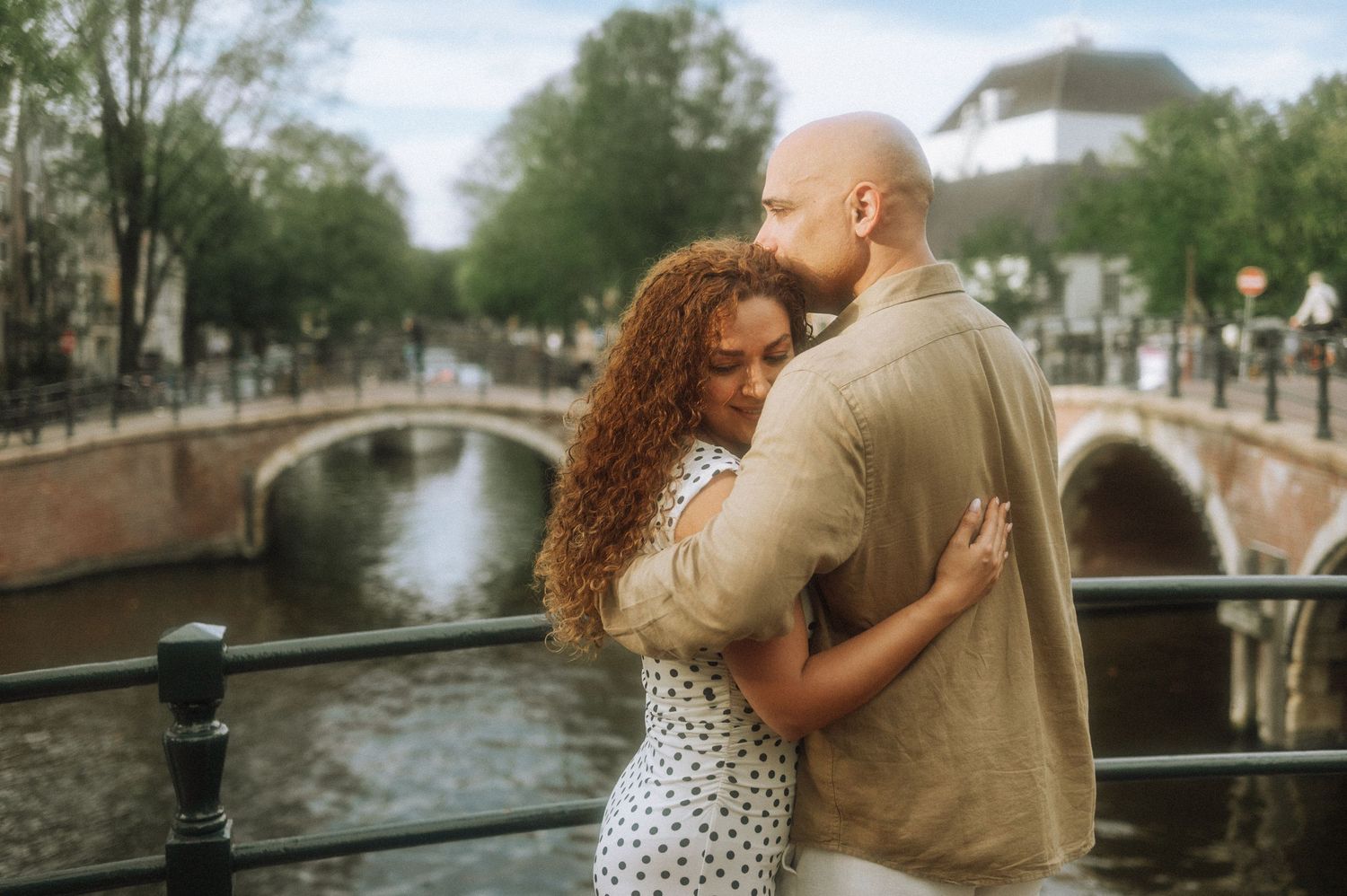 Romantic couple embracing on a canal bridge in a Amsterdam city, the woman with curly auburn hair wearing a polka dot dress.