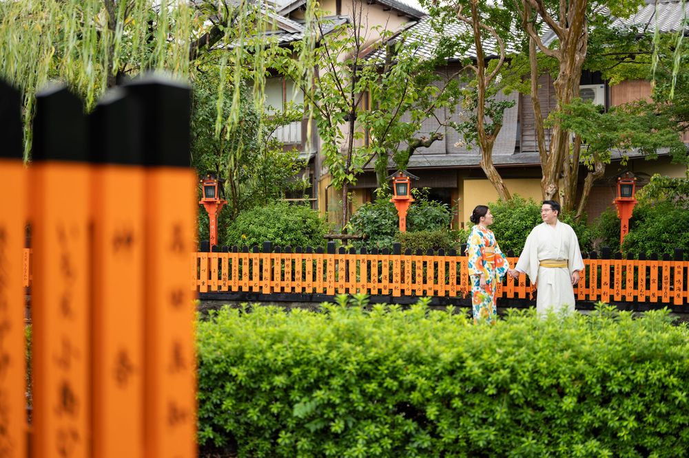 Two people in traditional Japanese kimono walk past an orange railing and lush green hedge in a peaceful Kyoto garden with willow trees.