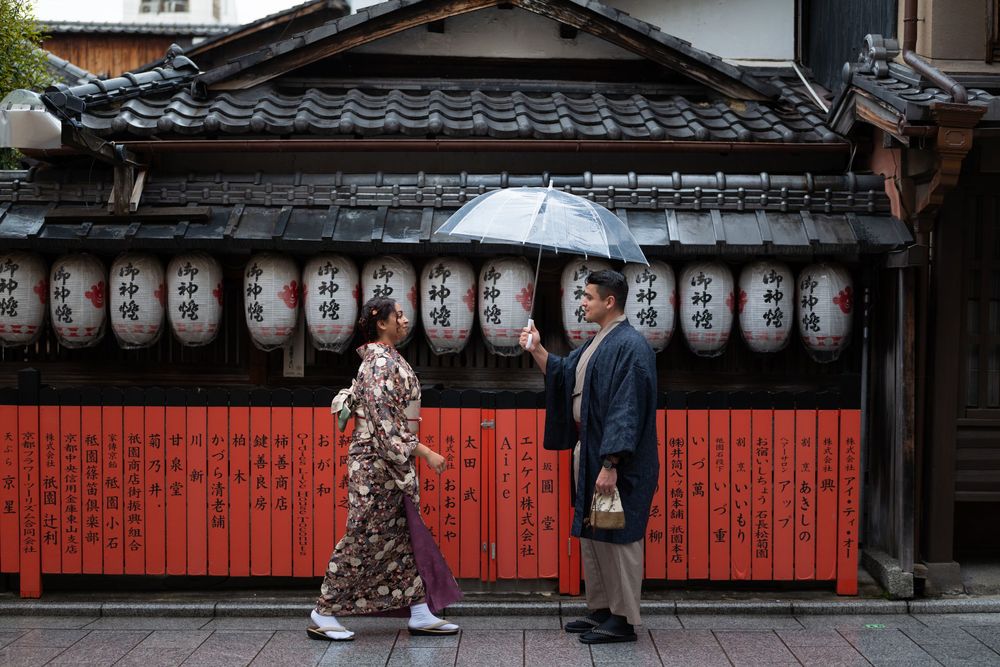 Couple in traditional Japanese kimono share an umbrella while standing before a red wooden wall with paper lanterns on a historic street.