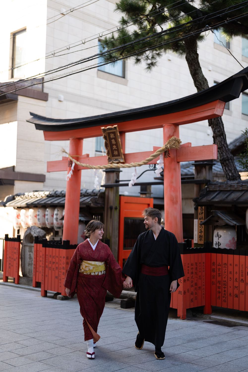 Couple in traditional Japanese kimono holding hands and walking past a red torii gate at a Shinto shrine in Kyoto.
